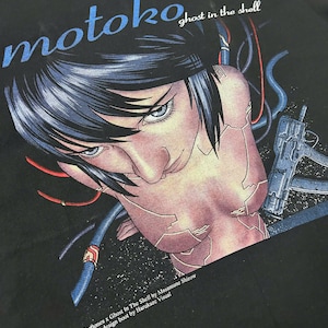 Puede incluir: Camiseta negra con un gráfico de Motoko de Ghost in the Shell, con texto azul y una ilustración detallada. El texto "motoko" y "ghost in the shell" son visibles. Diseño de Masamune Shirow y Harukaze Visual.