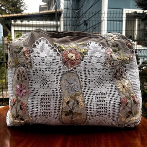 Könnte beinhalten: Eine handgefertigte, dekorative Tasche mit Patchwork-Design. Die Tasche hat eine Kombination aus grauem Stoff und weißer Spitze, verziert mit gestickten floralen Akzenten in Rosa, Gelb und Blau. Die Tasche steht auf einer Holzoberfläche.
