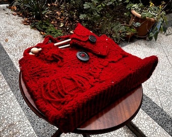May include: A handmade, red knitted bag with a flap and two large black buttons. The bag has a textured pattern and includes wooden knitting needles. It is sitting on a small, round wooden table.