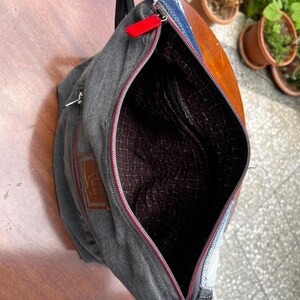 May include: A gray denim crossbody bag with a red zipper and a brown interior. The bag has a black strap and a small brown patch with text. The bag is open, revealing the interior lining.
