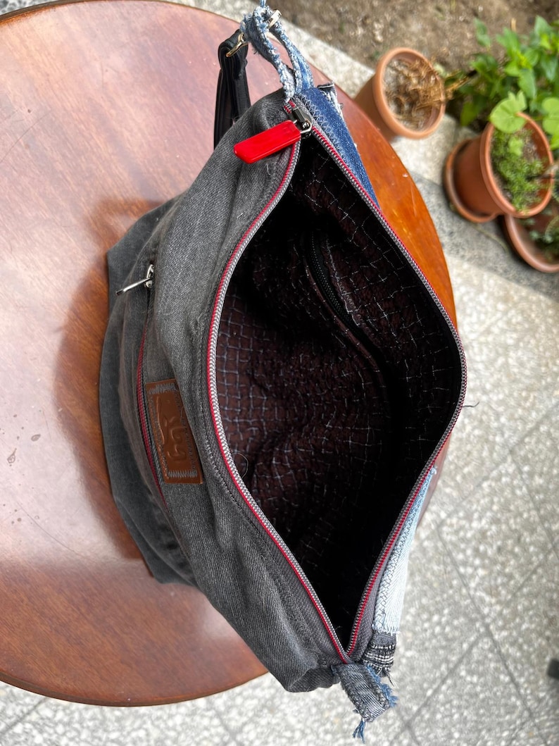 May include: A gray denim crossbody bag with a red zipper and a brown interior. The bag has a black strap and a small brown patch with text. The bag is open, revealing the interior lining.