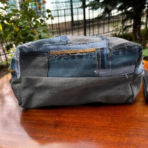 May include: A rectangular denim patchwork bag with a black handle. The bag is made from various shades of blue and gray denim, stitched together in a patchwork design. The bag is sitting on a wooden surface.