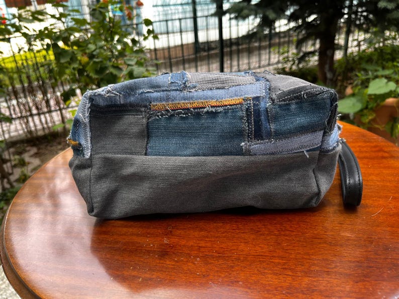 May include: A rectangular denim patchwork bag with a black handle. The bag is made from various shades of blue and gray denim, stitched together in a patchwork design. The bag is sitting on a wooden surface.