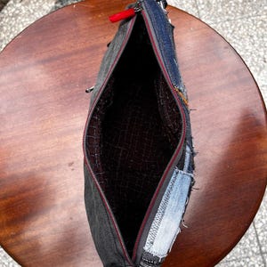 May include: Open denim clutch with a red zipper pull. The bag is made from repurposed denim patches in various shades of blue and gray. The interior lining is a dark patterned fabric.