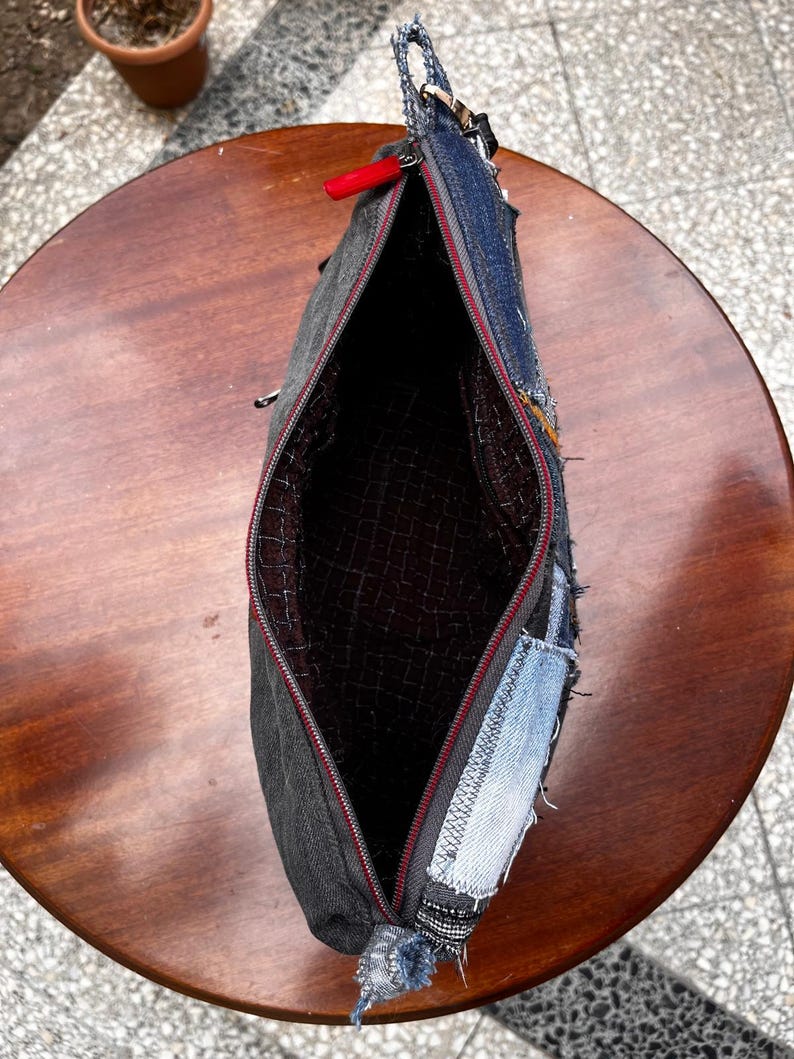 May include: Open denim clutch with a red zipper pull. The bag is made from repurposed denim patches in various shades of blue and gray. The interior lining is a dark patterned fabric.