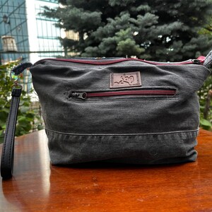 May include: A gray denim clutch bag with a red zipper and a leather wrist strap. The bag has a rectangular shape with a small leather patch on the front. The background includes greenery and a building.