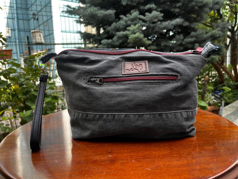 May include: A gray denim clutch bag with a red zipper and a leather wrist strap. The bag has a rectangular shape with a small leather patch on the front. The background includes greenery and a building.