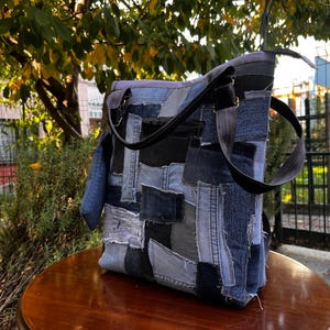 Op de afbeelding: Een patchwork denim tote bag met een zwarte band. De tas is gemaakt van verschillende tinten blauwe denim, waardoor een getextureerde, veelkleurige uitstraling ontstaat. De tas staat op een ronde houten tafel, met een vage achtergrond van bladeren en een gebouw.