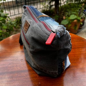 May include: A denim patchwork bag with a red zipper and a brown leather patch. The bag is made from various shades of blue and gray denim, with visible stitching and frayed edges. The bag is sitting on a wooden surface.