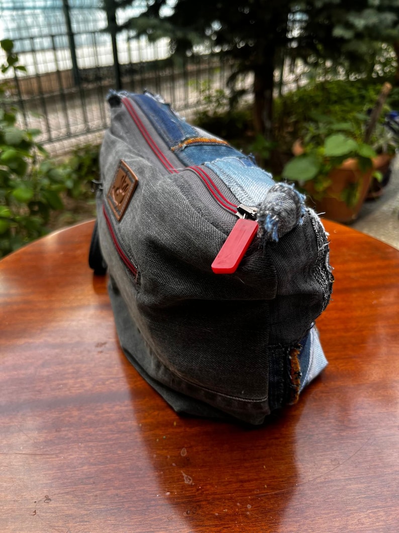 May include: A denim patchwork bag with a red zipper and a brown leather patch. The bag is made from various shades of blue and gray denim, with visible stitching and frayed edges. The bag is sitting on a wooden surface.