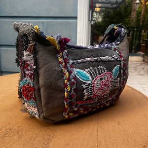 Könnte beinhalten: Eine graue Stoff-Umhängetasche mit bunten, gestickten Blumenmustern. Die Tasche hat ein Patchwork-Design mit roten, gelben und blauen Akzenten. Die Tasche hat einen geflochtenen Riemen und liegt auf einer braunen Oberfläche.