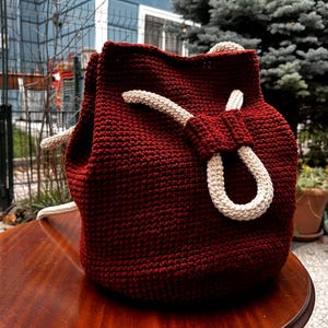 May include: A handmade, burgundy crochet drawstring bag with cream-colored rope details. The bag is sitting on a wooden table. The bag's texture is visible, and the cream-colored rope forms a loop closure.