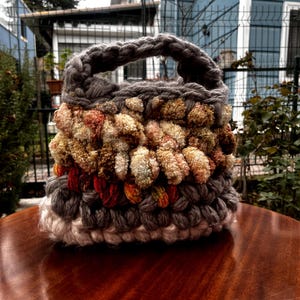 May include: A handmade, textured handbag with a braided handle. The bag features a mix of gray, beige, and red yarn, creating a unique, layered design. The bag is sitting on a wooden surface.