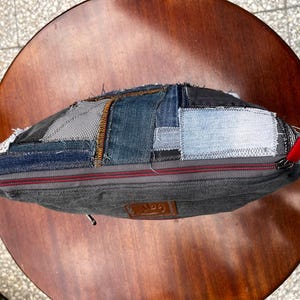 May include: A patchwork denim bag with a gray body and a red zipper pull. The bag is made from various denim patches in shades of blue and gray. The bag is sitting on a round wooden table.