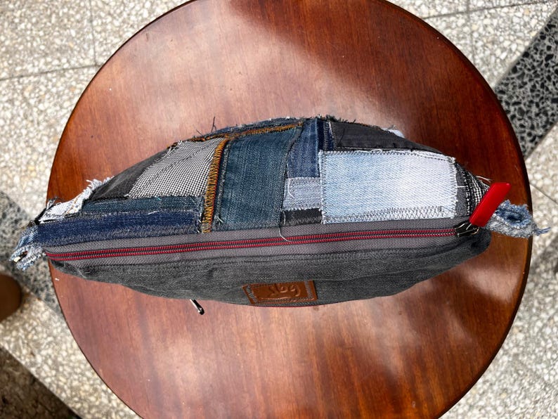 May include: A patchwork denim bag with a gray body and a red zipper pull. The bag is made from various denim patches in shades of blue and gray. The bag is sitting on a round wooden table.