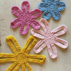 May include: Four handmade crochet flower appliques in bright colours. The set includes one yellow, one pink, one light pink, and one light blue flower. Each flower features a circular centre with six elongated, open-loop petals made of yarn.