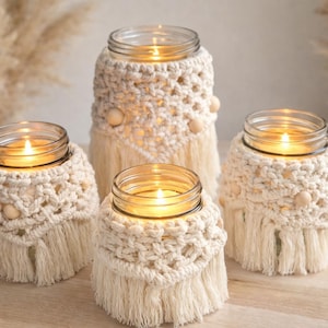 Pode incluir: Quatro velas acesas em potes de vidro, cada uma envolta em um design de macramê creme. O macramê apresenta um padrão tecido com contas de madeira e uma parte inferior com franjas. As velas estão em uma superfície de madeira clara, criando um ambiente acolhedor.