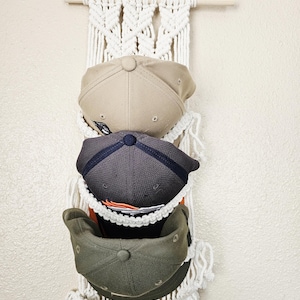 May include: A macrame hat organizer with three baseball caps. The caps are in shades of tan, gray, and olive green. The organizer is made of white macrame and has a wooden dowel at the top for hanging. The caps are displayed in pockets.