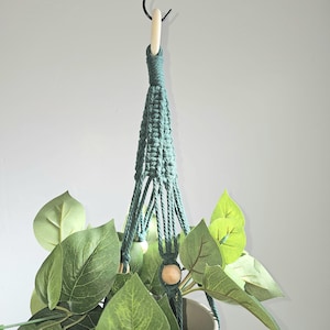 May include: A hanging macrame plant holder in teal green, suspended from a black hook. The macrame design cradles a white pot filled with lush green leafy plants. A wooden bead accents the design, adding a natural touch.