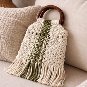 May include: Cream-colored macrame handbag with a central olive green stripe and fringe detail. The bag has a wooden handle and is displayed on a neutral-toned surface, showcasing a bohemian style.