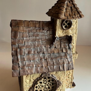 Może przedstawiać: Szczegółowy, miniaturowy domek z teksturowanym, brązowym dachem z gontów i małą wieżą. Dom posiada dekoracyjne okrągłe okna i beżowe, kamienne zewnętrzne, tworząc rustykalną estetykę.