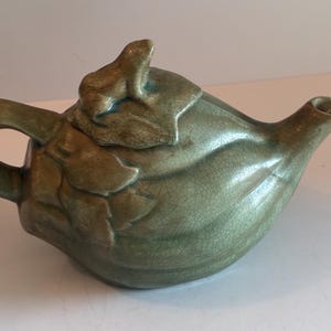 May include: A green ceramic teapot with a leaf design and a small frog on the lid. The teapot has a curved handle and a spout. The surface has a crackle glaze.