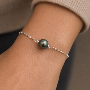 Puede incluir: Una delicada pulsera de plata con una sola perla gris oscuro iridiscente. La perla es el punto focal, conectada por una fina cadena. La pulsera se muestra en una muñeca, lo que sugiere una pieza de joyería.