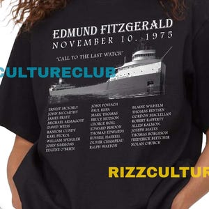 Op de afbeelding: Zwart t-shirt met de tekst "EDMUND FITZGERALD NOVEMBER 10, 1975", een scheepsafbeelding en een lijst met namen. Het shirt wordt gedragen met een spijkerbroekshort.