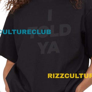 May include: Black t-shirt with the text "I TOLD YA" and "RIZZCULTURECLUB" in light blue. The model is wearing denim shorts. The background is a brick wall.