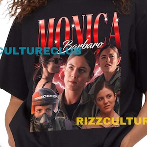 Puede incluir: Camiseta negra con un gráfico de Monica Barbaro y el texto "MONICA Barbaro" en rojo y blanco. La camiseta también tiene el texto "RIZZCULTURECLUB" en blanco. La modelo lleva pantalones cortos vaqueros.