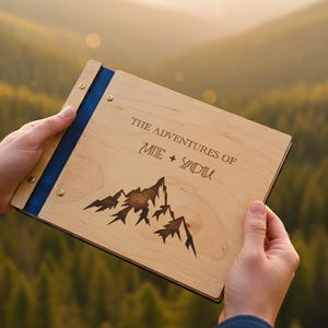 Könnte beinhalten: Ein Fotoalbum aus Holz mit dem Text "THE ADVENTURES OF ME + YOU" und einer Berg-Grafik. Das Album hat einen blauen Buchrücken und wird von zwei Händen vor einem verschwommenen Waldbild gehalten.