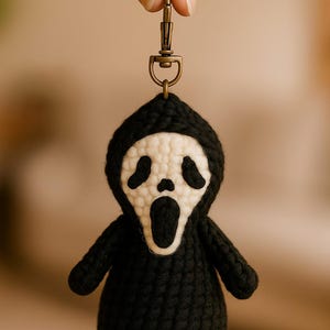 May include: A black and white crocheted keychain featuring a Ghostface mask design. The keychain has a black hooded robe and a white mask with black eye holes and an open mouth. The keychain is attached to a bronze-coloured clasp.
