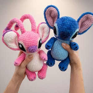 May include: Two handmade crochet plush toys: one pink with large ears and the other blue with large ears. Both toys have black eyes and are held up in front of a light background. The pink toy has purple accents, and the blue toy has pink accents.