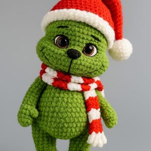 Könnte beinhalten: Eine grüne gehäkelte Kreatur mit großen braunen Augen und einer schwarzen Nase, die eine rot-weiße gehäkelte Weihnachtsmannmütze und einen passenden rot-weiß gestreiften Schal trägt. Die Figur hat einen runden Körper, kurze Arme und zwei Füße.