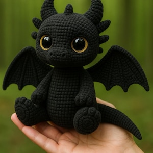 May include: A black crocheted dragon with large, golden-yellow eyes and small, pointed horns. The dragon has small wings and is sitting with its legs outstretched. The dragon is a handmade item.
