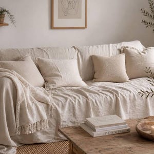 May include: A beige linen sofa with matching cushions and a fringed throw. A wooden coffee table holds books, a wooden bowl with a candle, and a vase with greenery. A small shelf with a potted plant is visible.