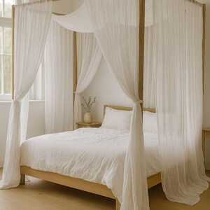 May include: A wooden four-poster bed with white bedding and sheer white curtains. The bed frame is light-colored wood, and the curtains are draped around the frame. The room has a neutral color palette with a wooden floor.