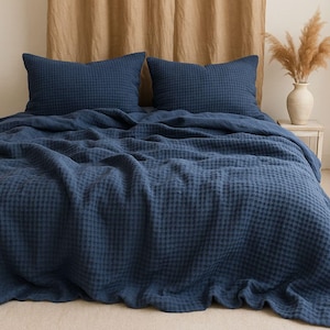 May include: A navy blue waffle-weave bed set, including two square pillowcases and a matching blanket. The bedding is arranged on a bed with a neutral-coloured backdrop and a vase of dried flowers.