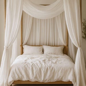 May include: A wooden four-poster bed draped with white sheer curtains. The bed is made with white linens and pillows. The room has a neutral color palette, with a beige rug and light-colored walls. The brand name FABTEXX is visible in the top right corner.