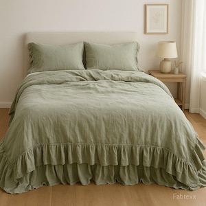 May include: Sage green linen bedding set on a bed. The set includes a duvet cover, two pillowcases, and a bed skirt with a ruffled edge. The bed is against a neutral-colored headboard. A small side table with a lamp and a framed picture are visible.