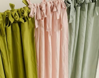 May include: Three linen curtains in shades of green, pink, and gray are displayed. Each curtain is suspended from a rod with fabric ties. The curtains are made of a soft, flowing material, and the colors are muted and natural.