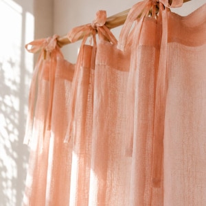 May include: A set of sheer, light peach-colored curtains hanging from a wooden rod. The curtains feature a textured, semi-transparent fabric and are attached to the rod with long, tied fabric ribbons at the top, creating a soft, draped effect.
