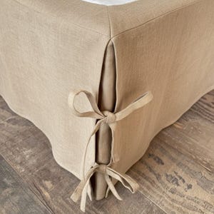 May include: Beige linen bed skirt with a tailored design. The skirt features a clean, simple aesthetic with corner pleats and decorative tie closures. The neutral color complements the white bedding.