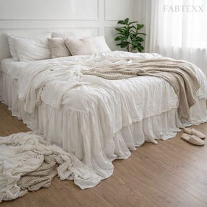 May include: A white bed with a ruffled bed skirt and matching bedding. Beige throw blanket and pillows add texture. The bed is in a bright room with a plant and a window.