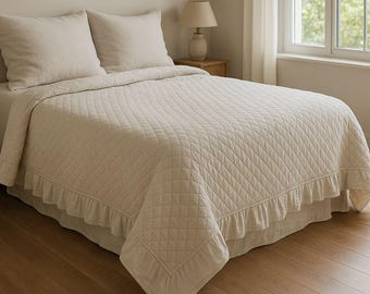 Könnte beinhalten: Eine cremefarbene Tagesdecke mit einem gesteppten Rautenmuster und Rüschenkanten. Zwei passende Kissen liegen auf dem Bett. Das Bett steht in einem hellen Raum mit Holzboden und Fenster.