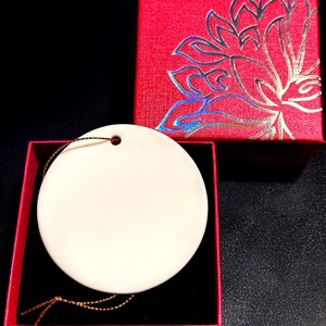 Op de afbeelding: Een ronde, cr&egrave;mekleurige ornament met een gouden draad, gepresenteerd in een open rode geschenkdoos met een zwart interieur. Een gesloten rode doos met een bloemmotief is ook zichtbaar.