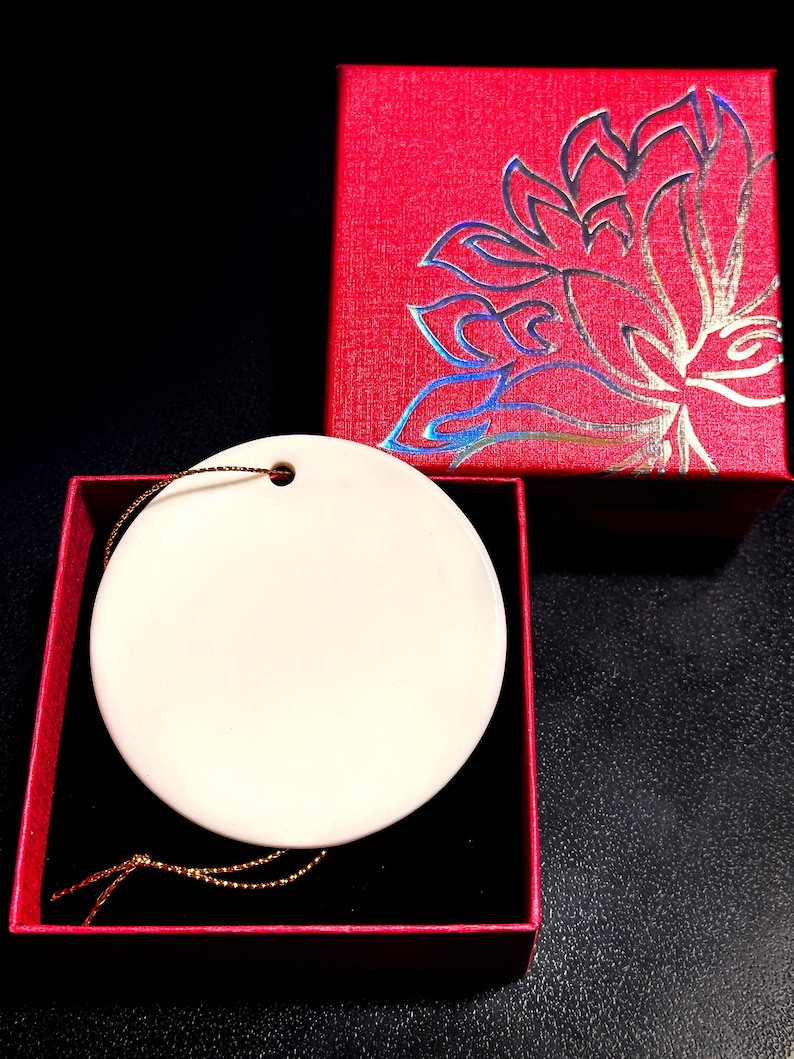 Op de afbeelding: Een ronde, cr&egrave;mekleurige ornament met een gouden draad, gepresenteerd in een open rode geschenkdoos met een zwart interieur. Een gesloten rode doos met een bloemmotief is ook zichtbaar.