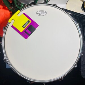 以下が含まれることがあります： 白いドラムヘッド、シルバーのリム、そして「UT CANOPUS」の文字。 「FUNNYBEADZ」の文字が入った黄色と紫のフロッピーディスクがドラムヘッドに取り付けられています。
