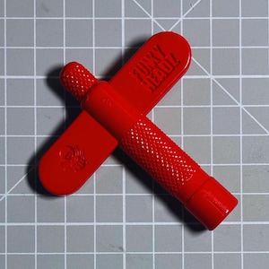 Può includere: Strumento rosso brillante con la scritta "FUNKY HEADZ" e un piccolo emblema. Lo strumento ha un'impugnatura strutturata e un design a forma di croce. Lo strumento è su una superficie a griglia grigia.