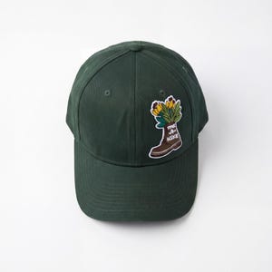 Könnte beinhalten: Dunkelgrüne Baseballkappe mit gebogenem Schirm. Die Kappe hat einen Aufnäher mit einem braunen Wanderstiefel mit Blumen und dem Text "Take A Hike". Der Hut ist aus weichem Material.